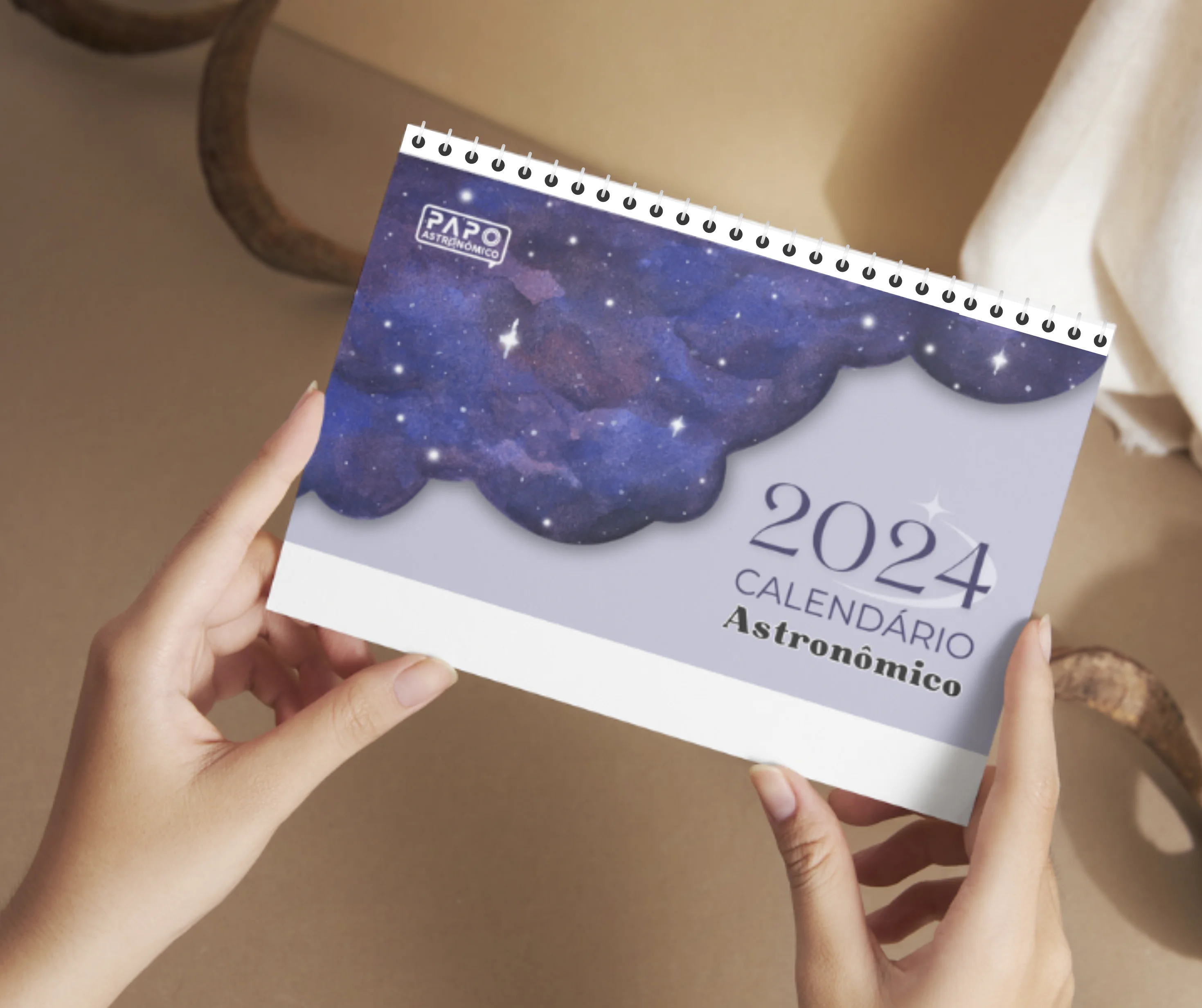 Mãos femininas seguram um calendário Astronômico do ano de 2024, ao fundo vemos parcialmente os detalhes de um cômodo da casa em tons de bege claro.